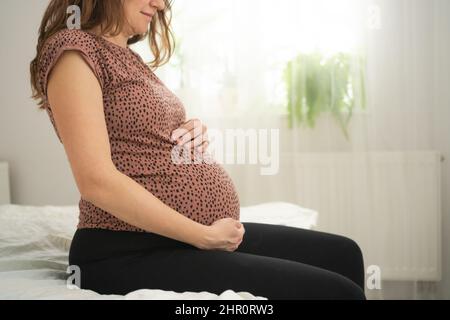 Une femme enceinte mignonne s'assoit dans le lit et tient sa main sur son ventre. Le concept d'une grossesse heureuse en prévision d'un bébé. 9 mois d'attente pour un bébé. Banque D'Images