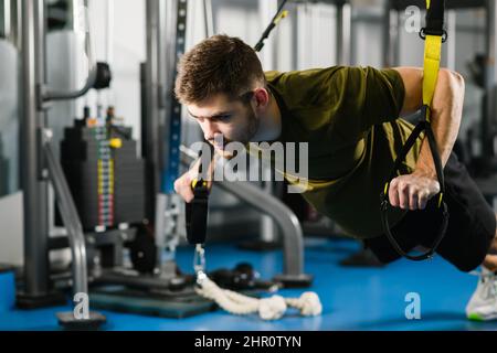 Homme fort dans les vêtements de sport faisant des push-up avec trx de forme physique. Jeune homme en forme à la main s'entraîner dans la salle de gym, un style de vie sain, un équipement moderne de croisement Banque D'Images