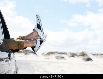 Faire une pause pour étirer mes jambes. Image rognée de deux pieds portant des baskets sortant d'une fenêtre de voiture garée avec des dunes en arrière-plan. Banque D'Images