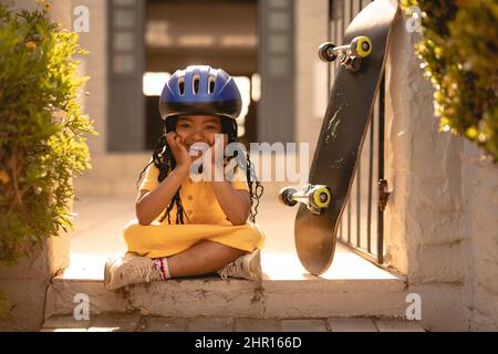 Portrait d'une jeune fille afro-américaine joyeuse portant un casque assis à pieds croisés par un skateboard à la porte Banque D'Images