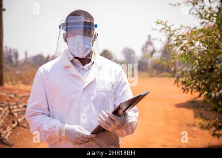 Portrait d'un médecin africain réussi, il porte un blouse de laboratoire, un masque chirurgical et un masque protecteur et tient un comprimé pour le travail Banque D'Images