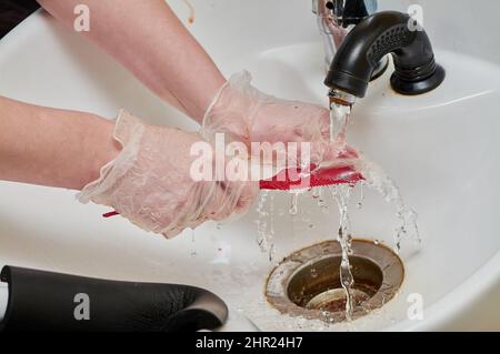 Les mains des femmes dans les gants de coloration des cheveux laver un peigne sous un jet d'eau dans un évier blanc Banque D'Images