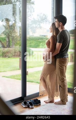 Jeune homme embrassant sa femme enceinte et regardant par la fenêtre Banque D'Images