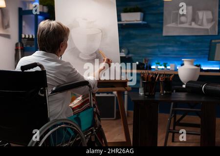 Femme plus âgée qui est paralysée ombre esquisse dans l'étude de lumière en classe de soirée sentiment inspiré dessin vase modèle sur papier. Professeur d'art à la retraite concentré en fauteuil roulant faisant des œuvres d'art créatives au crayon. Banque D'Images