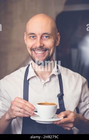 Homme souriant dans un tablier tenant une tasse avec un cappuccino de café. Le barista prépare du café chaud frais Banque D'Images