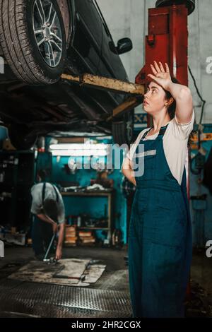 Égalité des sexes. Une jeune femme brune en uniforme se tient près de l'ascenseur avec la voiture, et regarde la roue, essuyant la sueur de son front. En t Banque D'Images