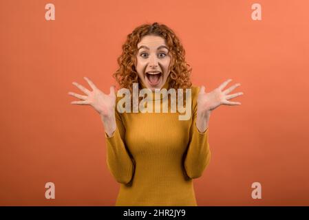 Portrait d'une jeune fille à tête rouge caucasienne attrayante, excitée et surprise dans un pull orange décontracté, isolé sur un fond orange studio. Banque D'Images