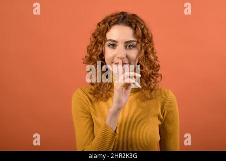 Jeune et attrayante fille caucasienne à tête rouge dans le cavalier orange montrant le geste de silence isolé sur fond orange studio. Banque D'Images