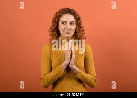 Jeune et attrayante rouge caucasienne fille en orange cavalier gestante avec les mains et plan de pensée isolé sur fond orange studio. Banque D'Images