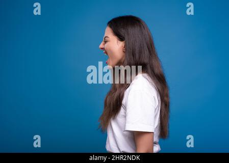 Jolie fille caucasienne ou arabe brune dans un t-shirt blanc est bouleversé et criant fortement isolé sur un fond bleu de studio. Banque D'Images