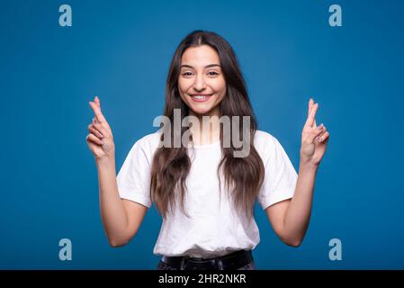 Le portrait d'une jeune fille brune blanche ou arabe heureuse et attrayante dans un t-shirt blanc a traversé les doigts et fait un souhait isolé sur un studio bleu Banque D'Images