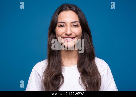 Portrait d'une jolie fille brune caucasienne ou arabe en t-shirt blanc isolé sur fond bleu de studio. Banque D'Images