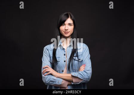 Portrait d'une jolie fille brune caucasienne en chemise avec bras croisés isolés sur fond noir de studio. Banque D'Images