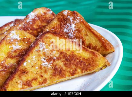 Une assiette de pain grillé. Cinq tranches de pain grillé se trouvent sur une assiette blanche, saupoudrées de sucre en poudre. La plaque est placée sur une nappe verte. Banque D'Images