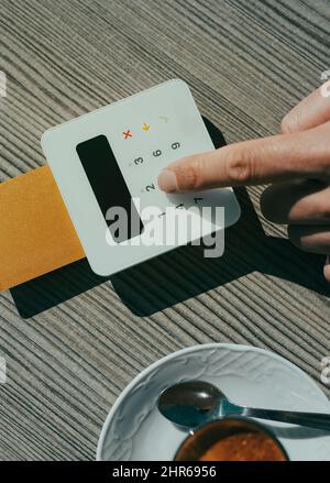 Vue en grand angle d'un jeune homme, assis à une table d'un café-terrasse, tout en insérant le code PIN de sa carte de crédit dans la manette d'un te de paiement Banque D'Images