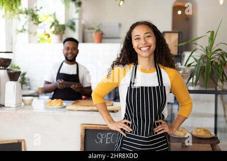 Portrait du jeune barista afro-américain souriant portant un tablier avec un collègue en arrière-plan Banque D'Images