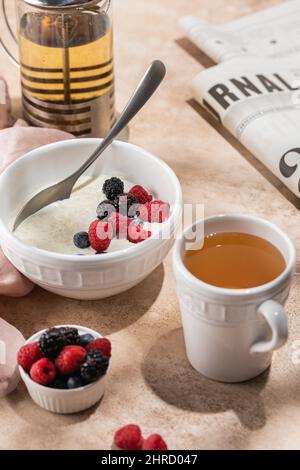 Petit déjeuner matin porridge de flocons d'avoine, céréales avec myrtilles fraîches et mûres dans un bol en céramique blanche, tasse de thé, journal, lumière dure, sélecti Banque D'Images