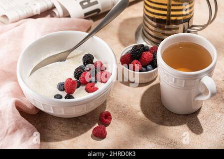 Petit déjeuner matin porridge de flocons d'avoine, céréales avec myrtilles fraîches et mûres dans un bol en céramique blanche, tasse de thé, journal, lumière dure, sélecti Banque D'Images