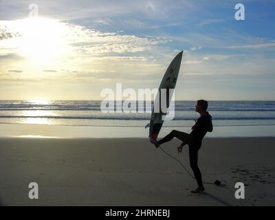 Surfeur adulte masculin au coucher du soleil faisant des exercices ou exposant sa planche de surf. Surfez et yoga ou étirez-vous après une séance de surf sur la plage. Surf gourou. Banque D'Images