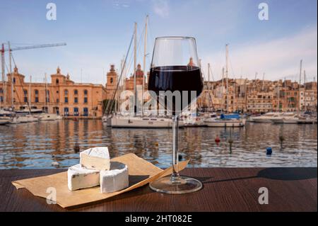 Verre de vin rouge avec fromage brie avec vue sur le port avec des bateaux et le centre historique de Birgu à Malte. Banque D'Images