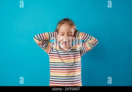 une petite fille couvre ses oreilles avec ses mains et hurle bruyamment avec sa bouche large ouverte sur un fond bleu isolé. Banque D'Images