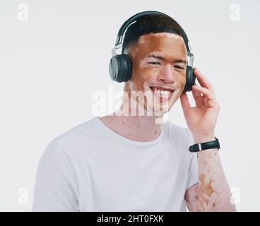 Lorsque les mots échouent, la musique parle. Portrait d'un beau jeune homme avec du vitiligo écoutant de la musique avec un casque sans fil sur fond blanc. Banque D'Images