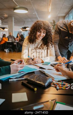Femme souriante aux cheveux bouclés étudiant avec des amis à la cafétéria Banque D'Images