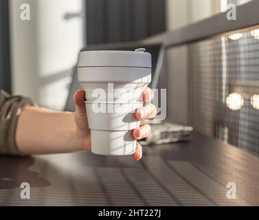 Femme gros plan tenant la tasse à emporter ECO pliable. Étudiant ou indépendant assis à table avec ordinateur portable et planificateur et boire du thé ou du café. Photo de haute qualité Banque D'Images