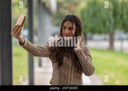 Jeune fille souriante regardant le smartphone, touchant et peignant les cheveux en utilisant le mobile comme un miroir dans la rue Banque D'Images