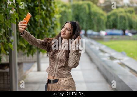 Jeune fille souriante regardant le smartphone, touchant et peignant les cheveux en utilisant le mobile comme un miroir dans la rue Banque D'Images