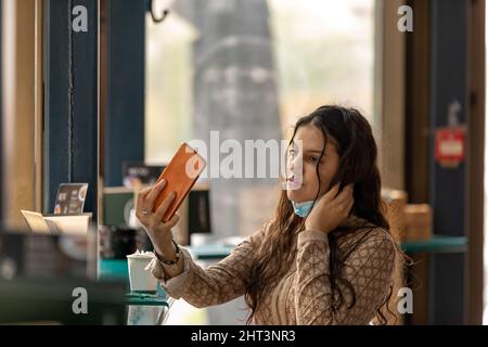 Un adolescent mignon portant un masque médical assis dans un café touchant et peignant les cheveux en utilisant un smartphone comme miroir Banque D'Images