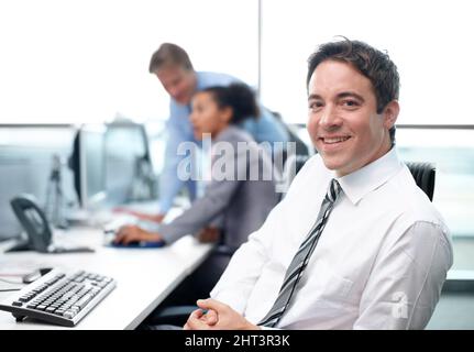 J'ai trouvé ma place au travail. Homme d'affaires confiant assis à son bureau avec ses collègues travaillant en arrière-plan - portrait. Banque D'Images