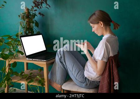 Sérieuse entreprise travail freelance fille femme en t-shirt blanc et jeans travaillant sur ordinateur portable et téléphone sur une table en bois contre le mur vert dans le bureau à la maison avec des plantes de maison Banque D'Images