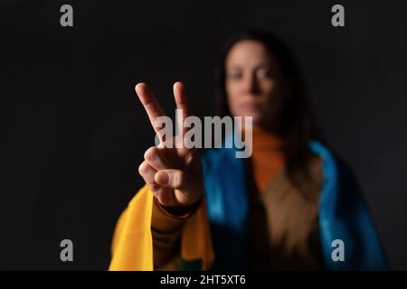 Triste femme couverte d'un drapeau de l'Ukraine montrant le signe de la paix. Concept de se tenir debout avec la nation ukrainienne en guerre avec la Russie. Banque D'Images