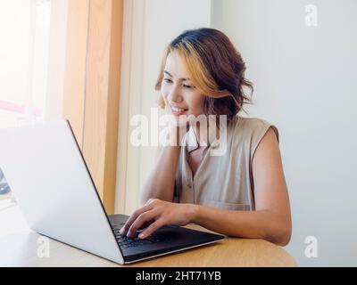 Femme asiatique, freelance avec cheveux courts tendance et regarder l'écran tout en travaillant avec un ordinateur portable sur un bureau rond en bois sur blanc W. Banque D'Images