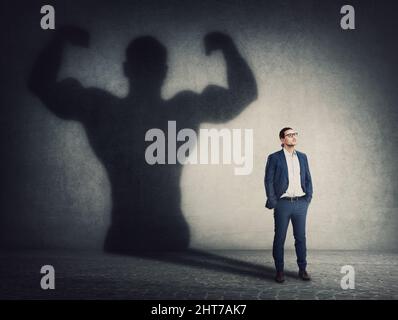 Un homme d'affaires confiant tient les mains dans ses poches tout en projetant une ombre puissante sur le mur derrière lui. La transformation de la personne d'affaires comme métaphore f Banque D'Images