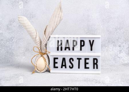 Traditionnel Wish Happy Easter sur un panneau lumineux. Oeuf enveloppé dans une serviette en lin à rayures nouée avec du jute en forme de lapin de Pâques avec des oreilles sur le gris clair Banque D'Images