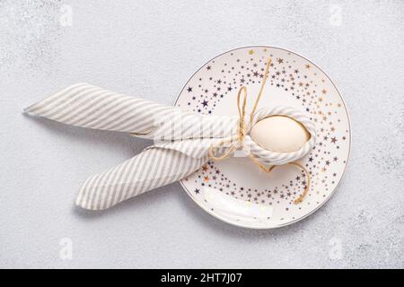 Oeuf enveloppé dans une serviette en lin à rayures nouée avec du jute en forme lapin de Pâques avec des oreilles sur l'assiette, fond gris clair. Idée de la mise en place de la table de Pâques Banque D'Images