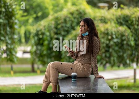 Belle jeune fille avec un smartphone et un café assis dans un parc et ayant une conversation téléphonique. Copier l'espace Banque D'Images