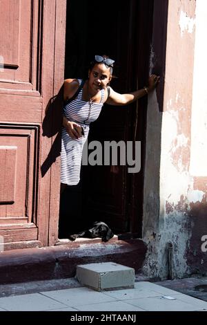 Une jeune femme tient son téléphone portable et se penche contre la porte tandis que son chien de compagnie se pose sur le sol à la porte de leur maison à la Havane, Cuba. Banque D'Images