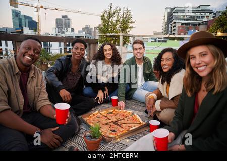 Groupe d'amis divers souriant à l'appareil photo avec des coupes rouges tout en étant assis sur un toit-terrasse dans la ville Banque D'Images
