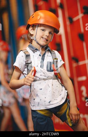 Happy School Boy à Red Helmet en cours d'escalade au centre d'escalade intérieur. Garçon souriant à l'appareil photo et tenant le pouce vers le haut Banque D'Images