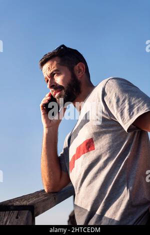 portrait vertical d'un beau jeune homme parlant sur son smartphone penché sur une rampe en bois avec le ciel bleu en arrière-plan, concept de loisir a Banque D'Images