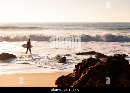 surfeur méconnaissable marchant le long du rivage de la plage avec sa planche sous son bras au coucher du soleil, concept de loisirs et de détente, copier l'espace pour le texte Banque D'Images