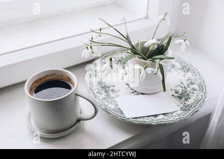 Scène de vie d'hiver, de printemps. Tasse de café sur le rebord de la fenêtre. Vase de fleurs de neige sur une assiette décorative verte vintager. Motif floral féminin Banque D'Images