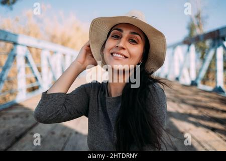 Portrait d'une femme hispanique souriante portant un chapeau assis sur un quai au bord de la rivière par une journée ensoleillée Banque D'Images