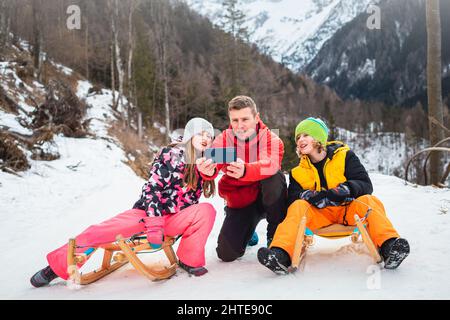 Père avec des enfants prenant des selfies tout en étant assis sur des traîneaux en bois, avec un magnifique paysage de montagne en arrière-plan. Banque D'Images