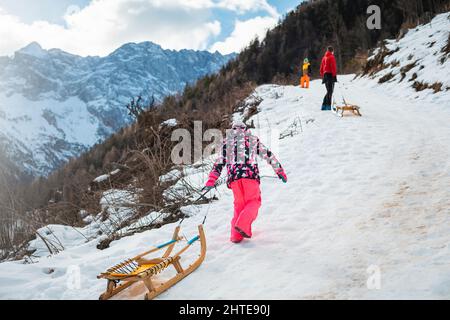 Fille en costume de neige coloré marchant derrière son père et son frère, allant en montée et tirant un traîneau en bois. Banque D'Images