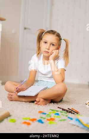 Photo verticale d'une petite fille primaire fatiguée qui écrit ses devoirs dans un ordinateur portable assis sur le sol à la maison en regardant l'appareil photo. Banque D'Images