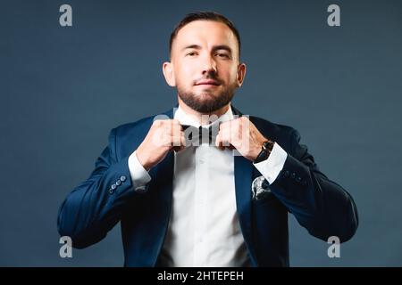 Le jeune homme d'affaires en costume ajuste son noeud papillon avec ses mains et regarde la caméra. Portrait de studio sur fond gris. Un homme dans un costume classique Banque D'Images